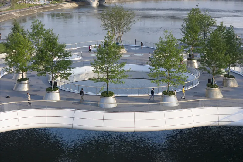 Circular plaza with trees emerging from conical planters overlooking the river with pedestrians strolling beneath