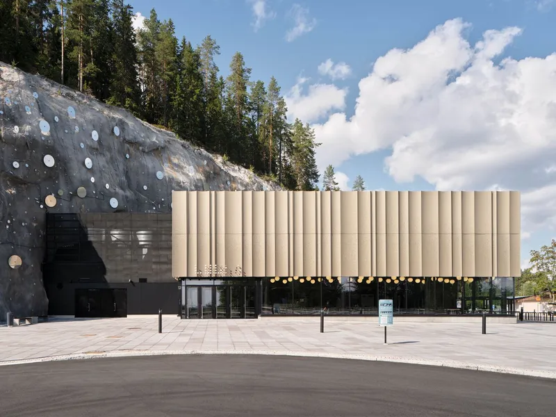 Plaza view showing ribbed facade panels beside a rock-cut excavation with circular insets under billowing clouds