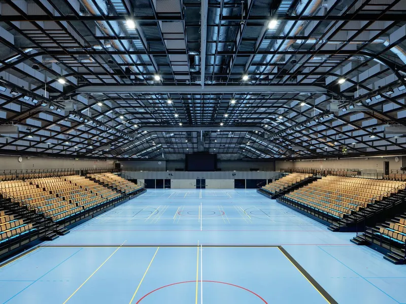 Sports hall interior with undulating steel truss roof and tiered seating overlooking blue court markings