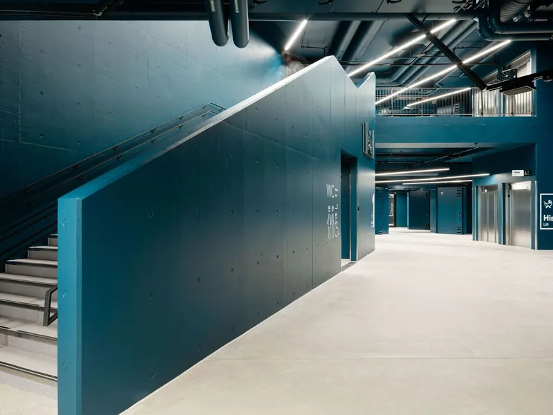 Interior lobby with teal concrete walls enclosing an escalator and stairs beneath exposed ceiling ducts