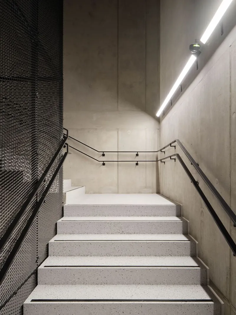 Concrete stairwell with metal mesh balustrade and continuous strip lighting along the upper wall corner
