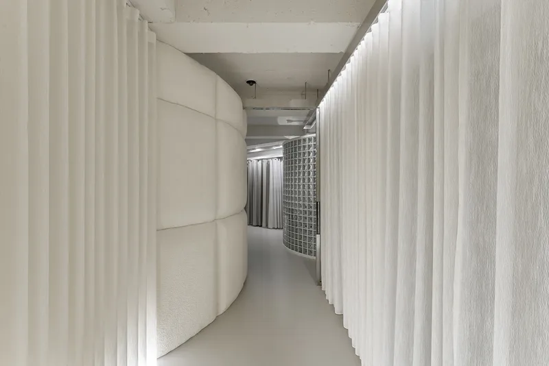 Narrow corridor defined by curved padded wall and white vertical curtains under concrete soffit