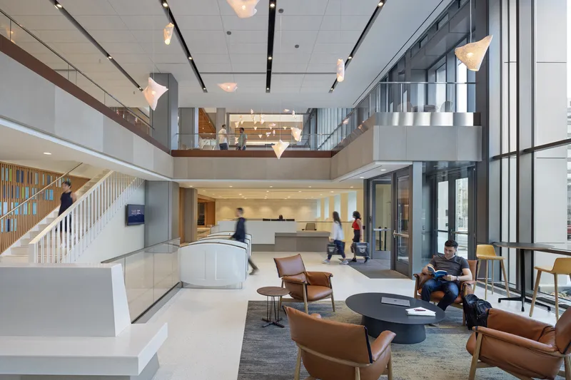 Double-height lobby with white staircase, suspended pendant lights, and people gathered around leather seating