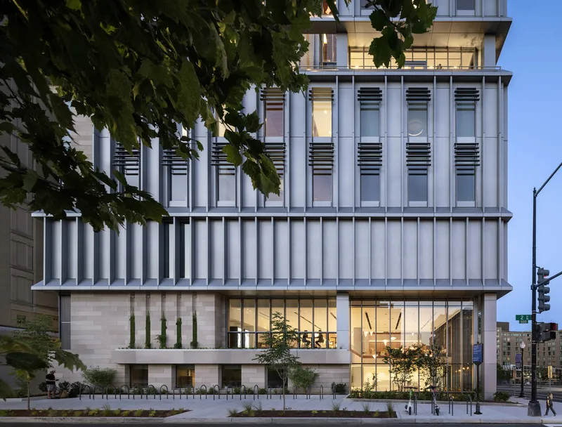 Street facade at dusk with illuminated lobby, vertical white cladding, and tree canopy in foreground