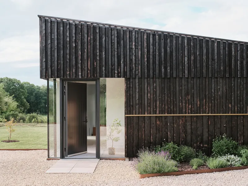 Charred timber facade with open folding glass doors revealing the white interior vestibule and gravel forecourt