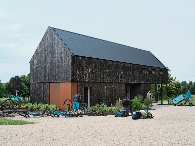 Charred timber house on gravel with family unloading bikes and camping gear in the forecourt