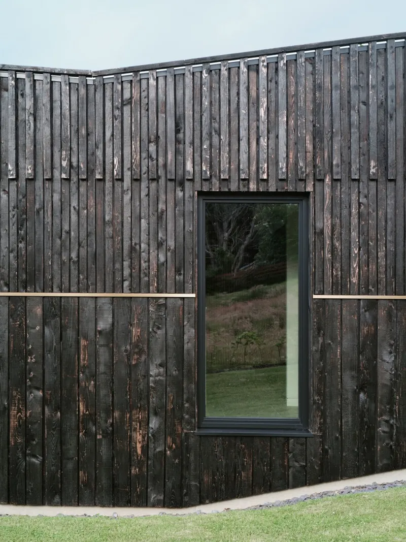 Tall window framed in charred vertical timber reflecting the lawn and trees at dusk