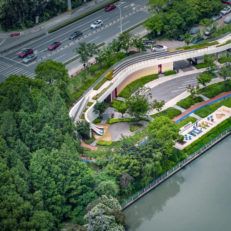 Aerial view of the curved riverside park with white terraces, planted beds, and playground between roadway and canal