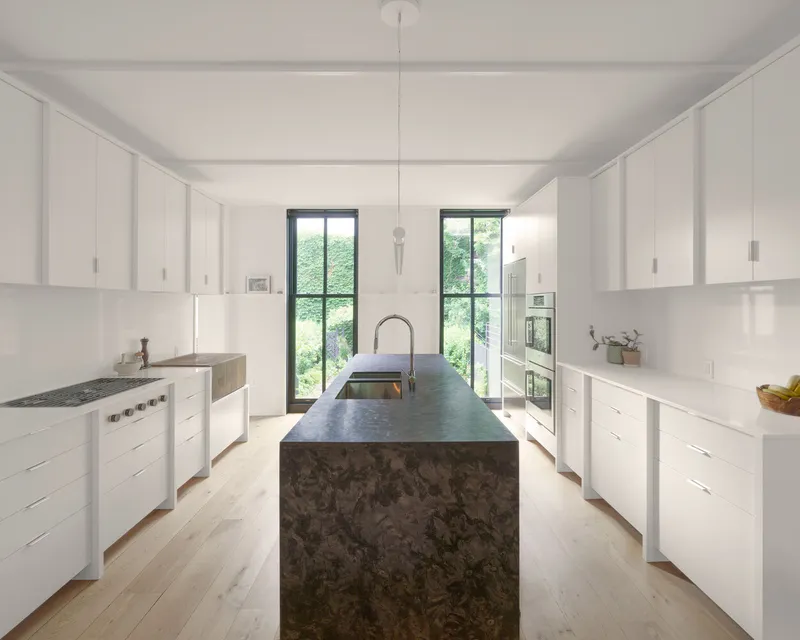 Galley kitchen with white cabinetry and dark stone island beneath twin windows facing greenery