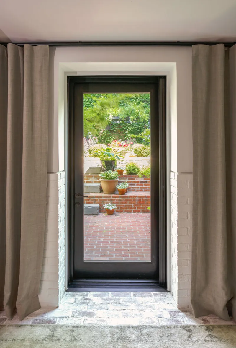 Black-framed glass door with linen curtains opening onto a brick courtyard with potted plants