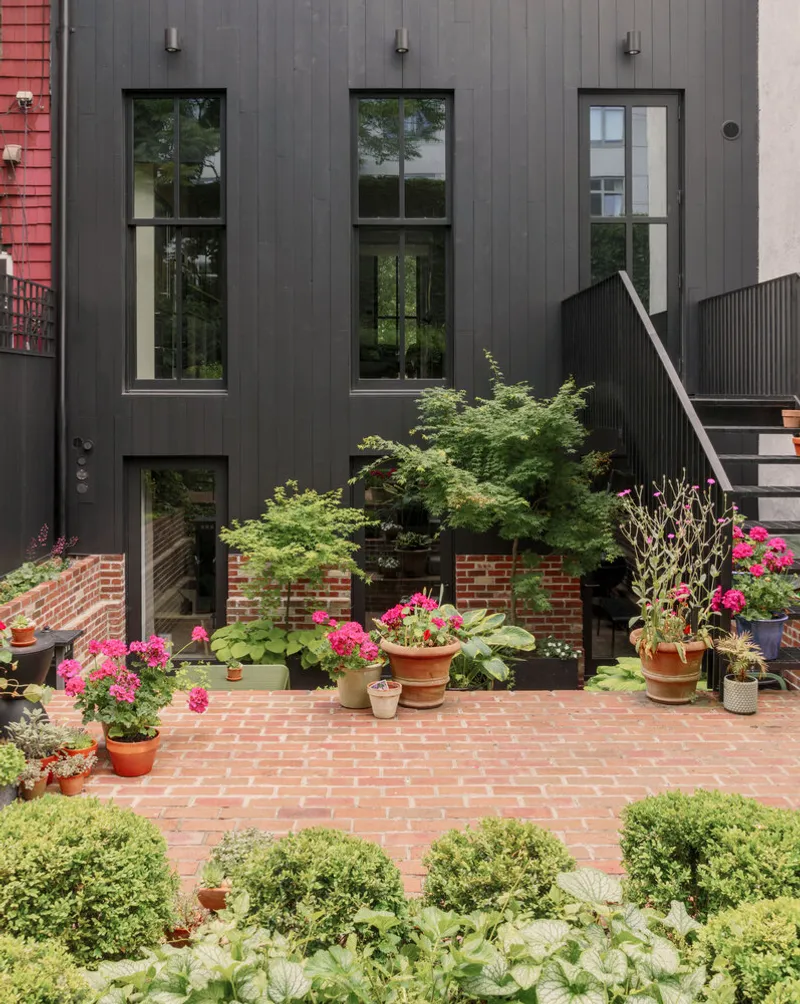 Dark vertical paneled facade with tall windows overlooking brick patio and potted flowering plants