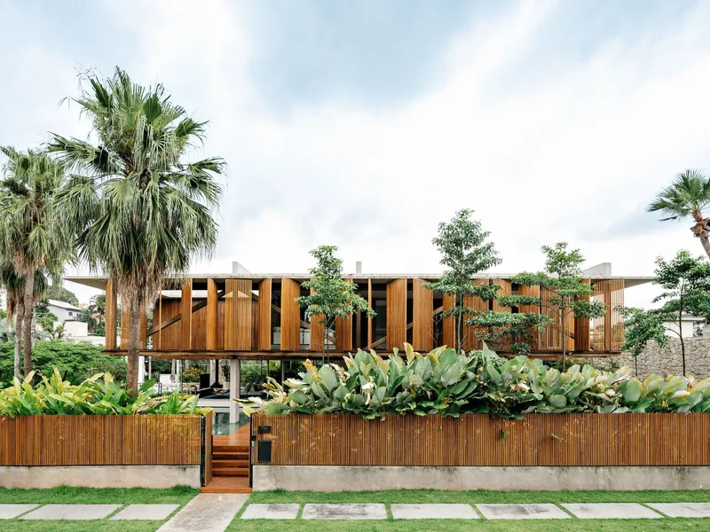 Street view of the elevated timber-clad volume on concrete columns with palm trees and bamboo fence