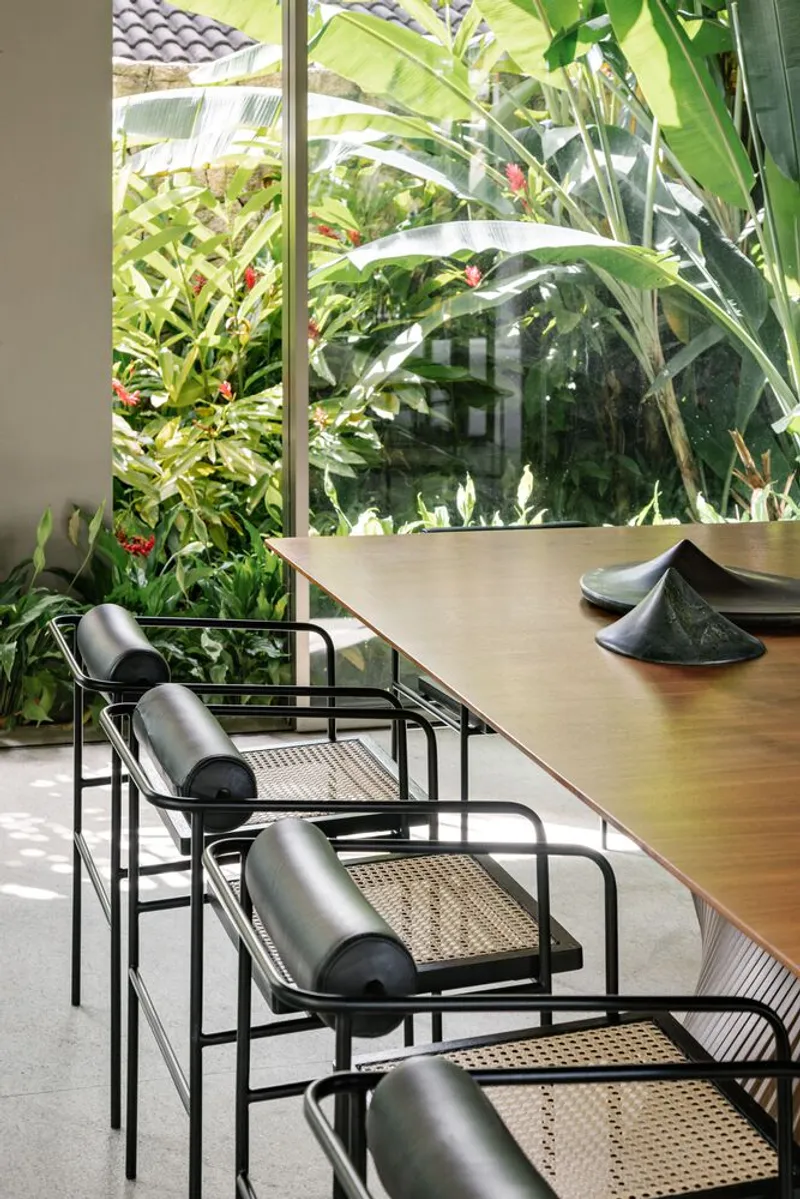 Dining table with black cane chairs facing floor-to-ceiling windows overlooking banana palms and tropical vegetation