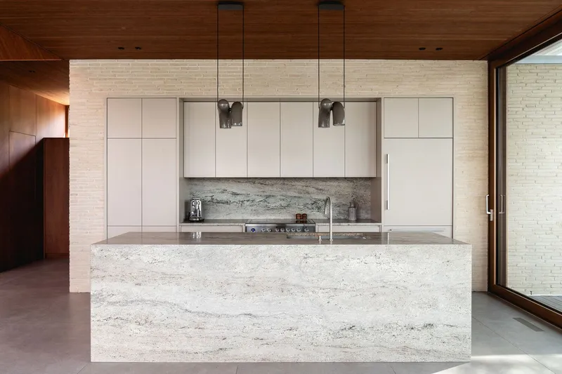 Kitchen with travertine island and white cabinetry against cream brick wall with pendant lights overhead