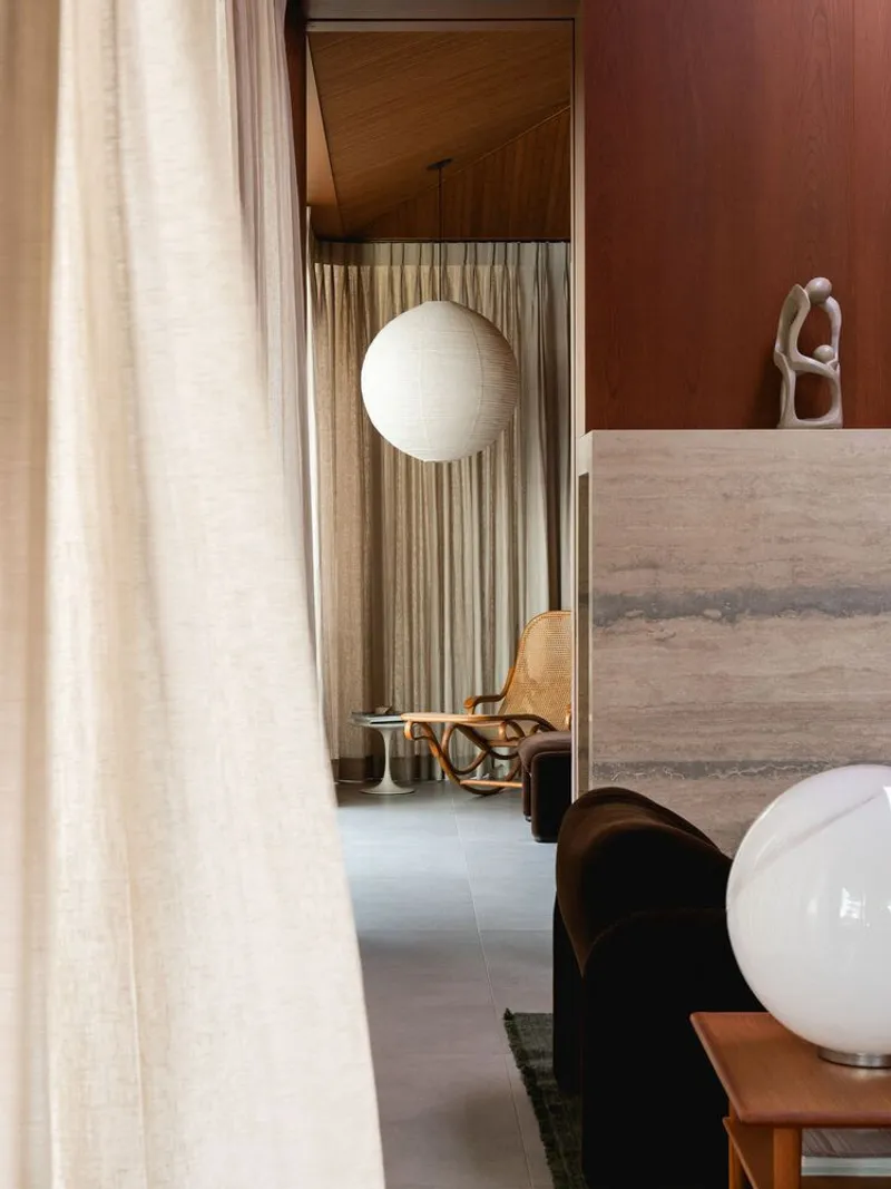 Living room corner with sheer curtain, spherical pendant light and travertine bench against wood-paneled wall