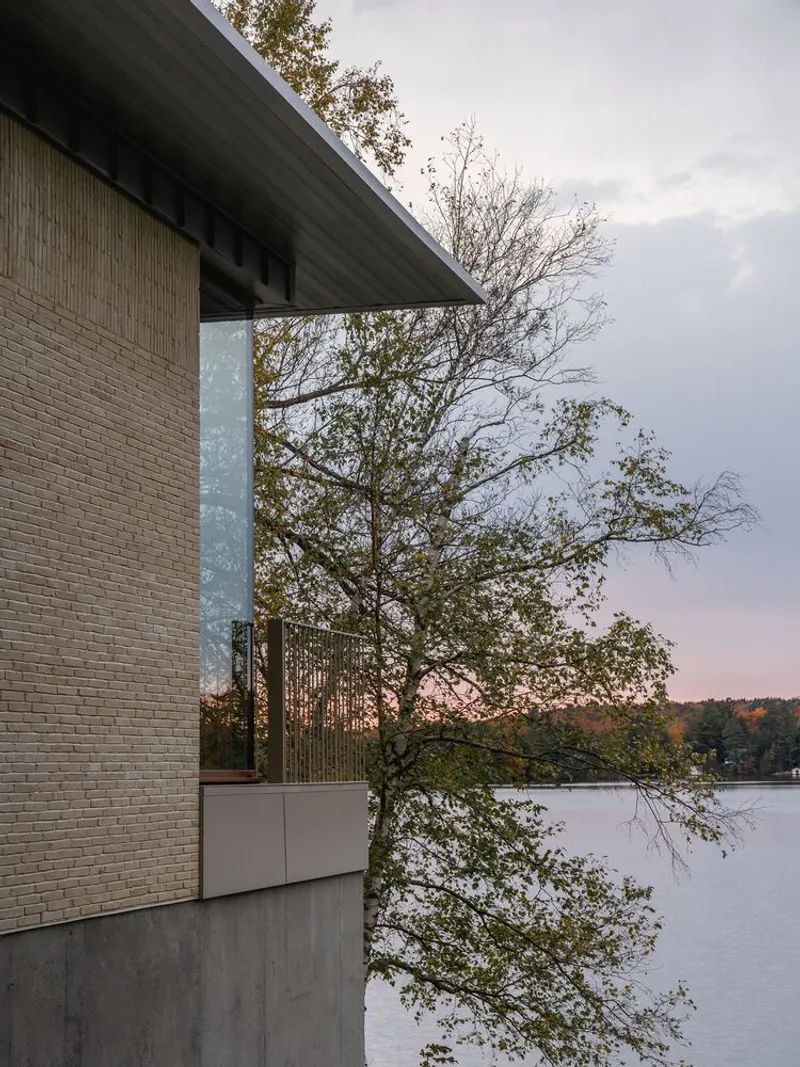 Exterior corner with textured brick wall, concrete overhang and balcony railing framed by bare trees and a lake