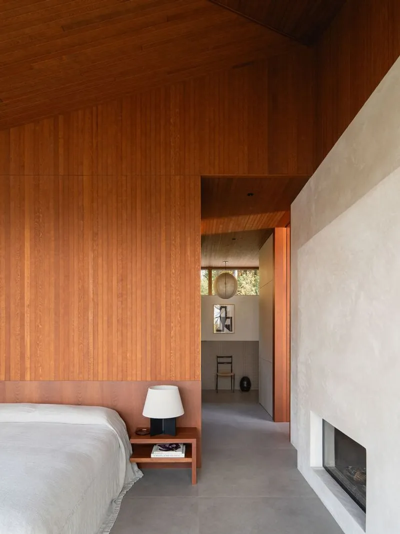 Bedroom with vertical wood-paneled walls and ceiling, concrete floor and white plaster fireplace wall with recessed opening