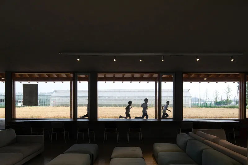 Interior lounge with timber-framed windows overlooking a playing field where children run past at midday