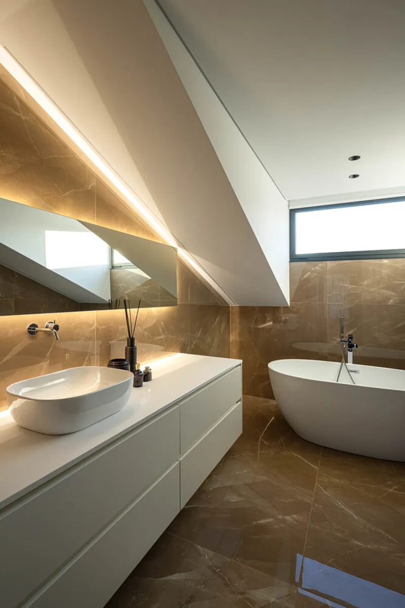 Bathroom with sloped ceiling, backlit mirror, white vanity, freestanding tub, and brown marble wall tiles
