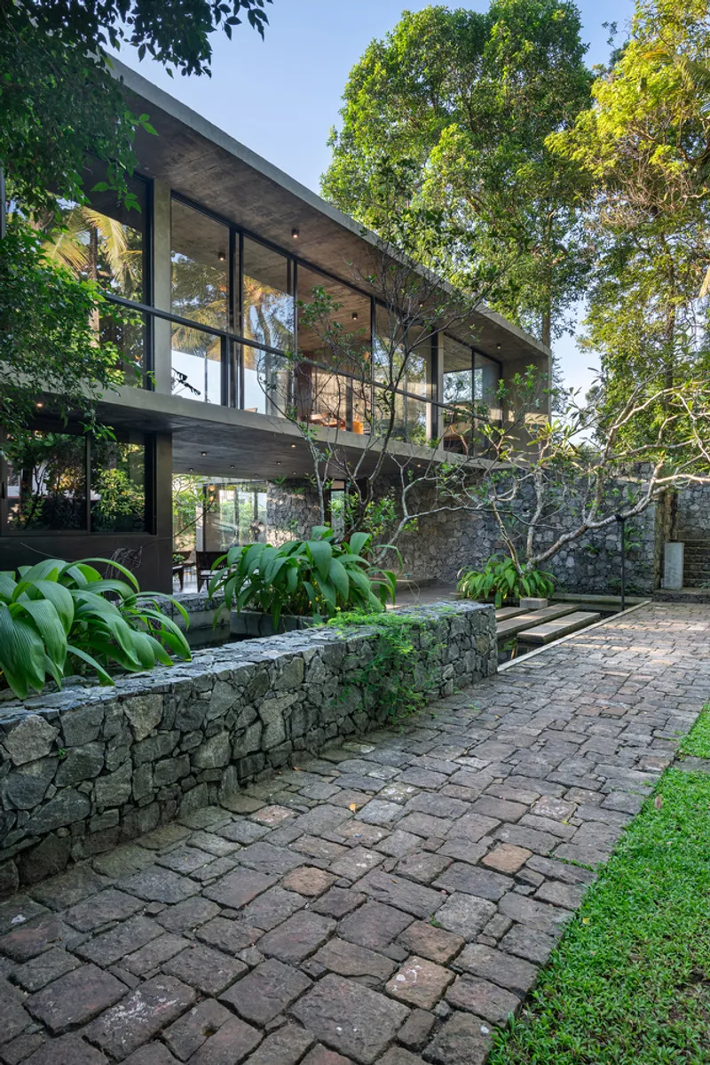 Gabion-wall planted beds and brick paving leading to the elevated glass volume under tree canopy