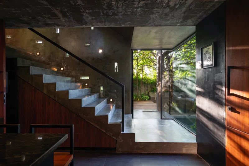 Concrete staircase with steel railing rising past a glazed corridor opening to the planted courtyard with dappled sunlight