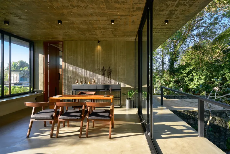 Dining area with timber table and exposed concrete ceiling opening to a terrace overlooking dense tree canopy