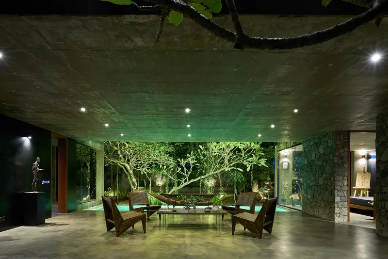 Covered terrace with board-formed concrete ceiling, wooden lounge chairs, and uplighting on trees at night