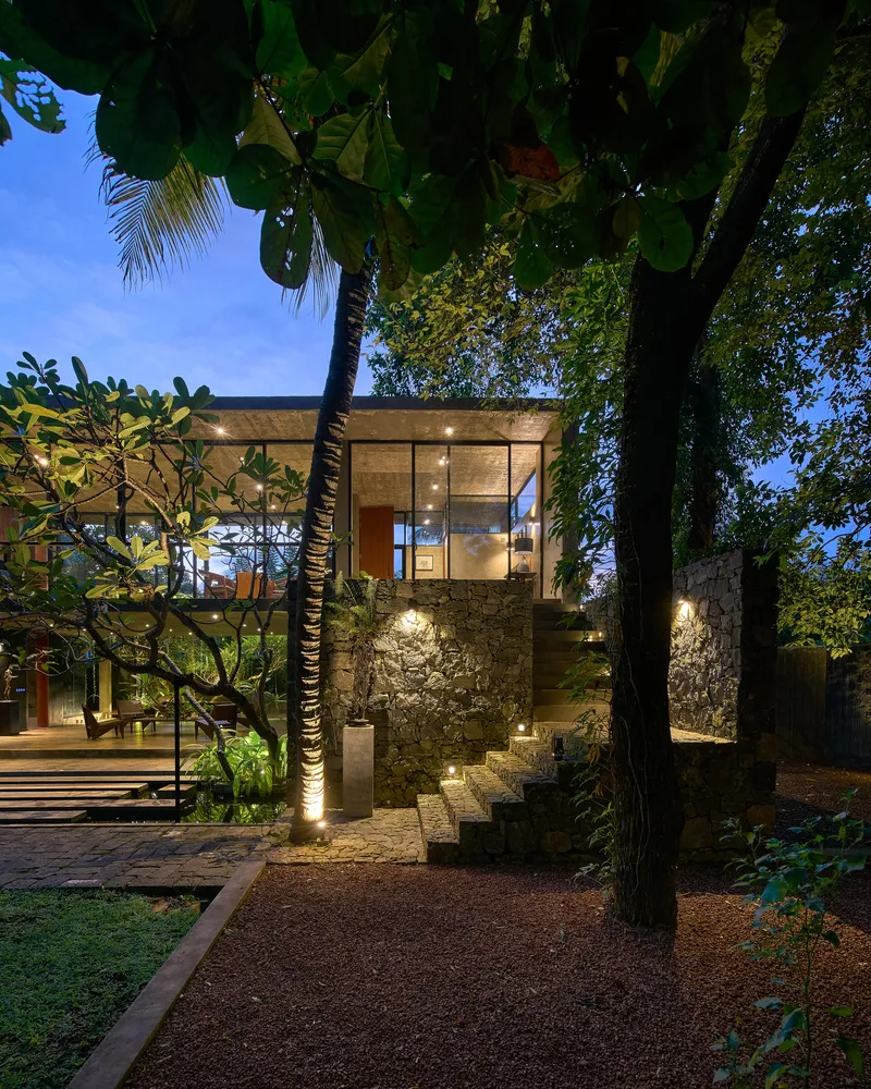 Illuminated glass pavilion on stone base framed by palm trees and tropical foliage at dusk