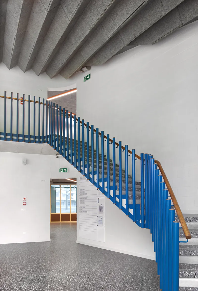 Curved staircase with blue vertical balustrades and timber handrail beneath angled concrete ceiling slabs