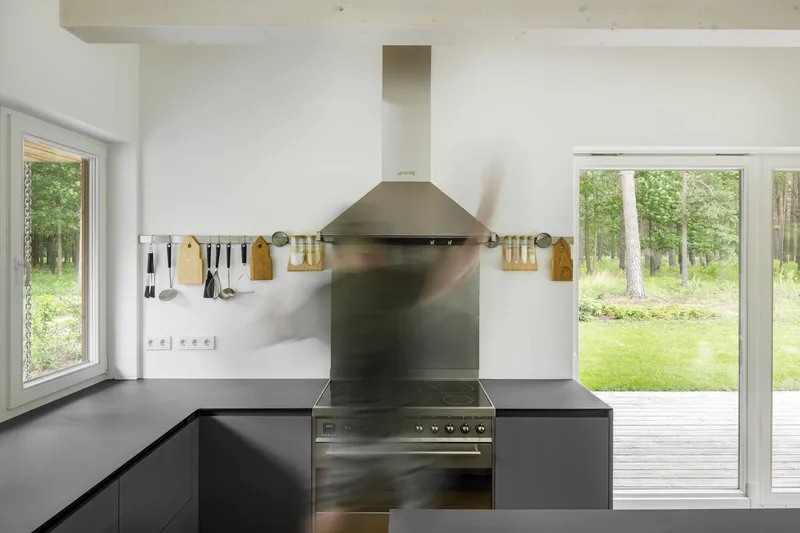 Kitchen with stainless steel range hood and black cabinets framed by windows overlooking a forest