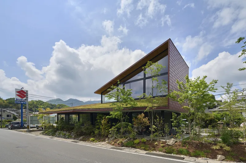 Street view of the building showing the tiled upper volume and planted landscaping under summer clouds