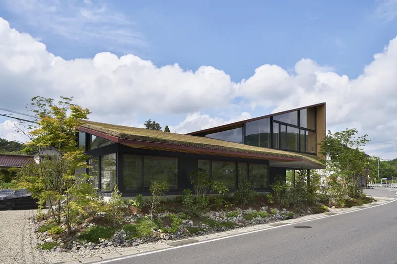 Street view of the sloping green roof and glass-walled facade with landscaped garden bed under cloudy skies