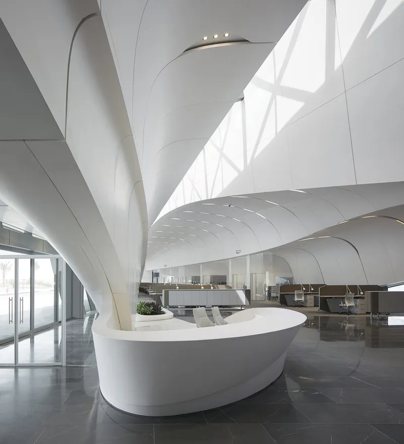 Reception area with curved white counter below branching columns and triangular glazed wall with natural light