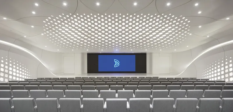 Auditorium with tiered seating facing a central screen beneath a perforated ceiling installation