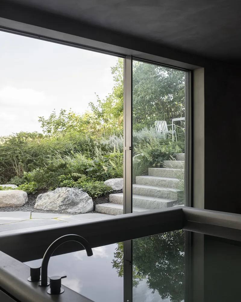 Interior view through floor-to-ceiling glass doors toward concrete garden steps and lush planting beds with stone boulders