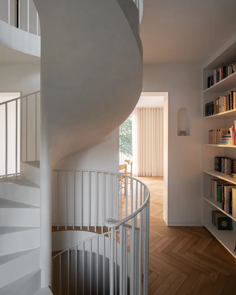 White spiral staircase with curved soffit rising through a herringbone oak floor and built-in bookshelves