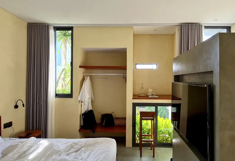 Bedroom interior with pale plywood walls, timber shelving, and tall windows framing palm fronds outside