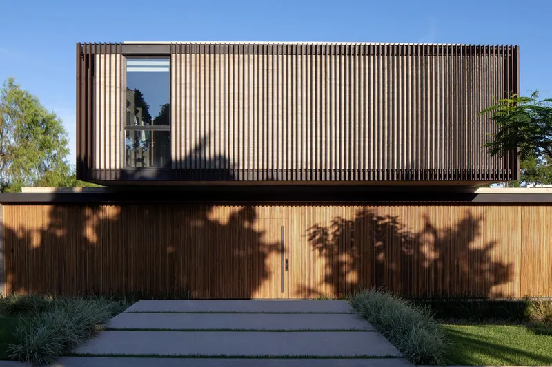 Street facade with vertical timber screening and concrete path casting tree shadows across the ground level