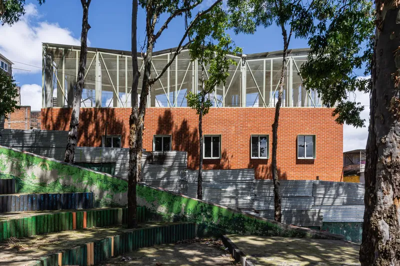 Layered facade with corrugated metal base, red brick midsection, and diagonal-braced glass pavilion above, framed by tall eucalyptus trees