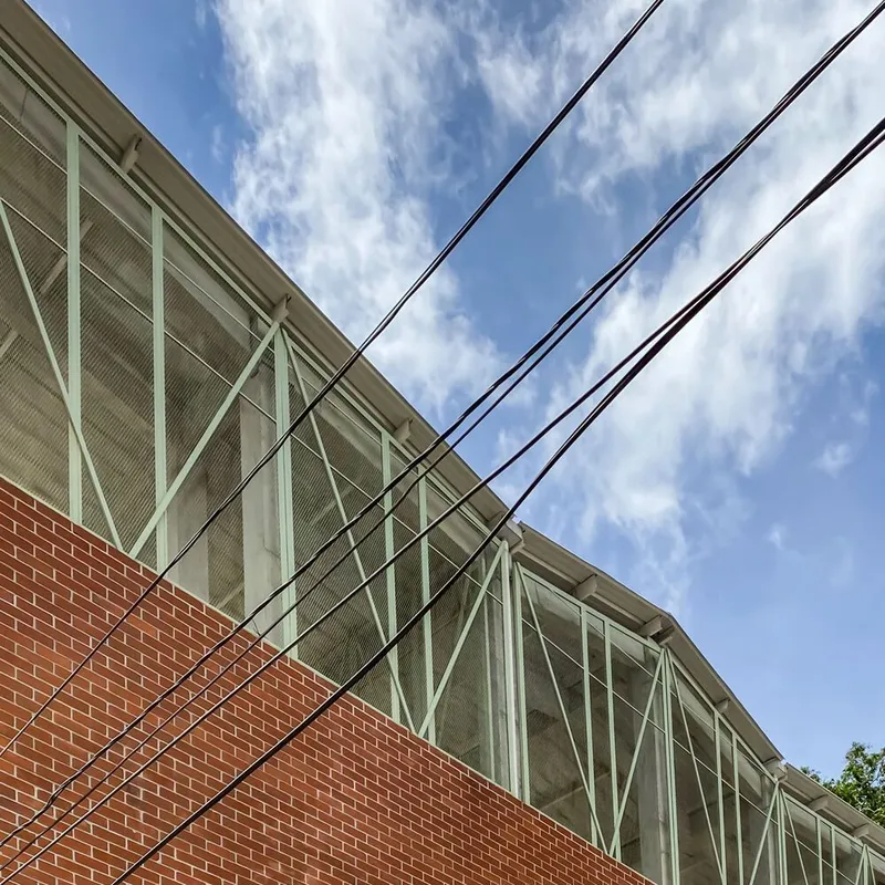 Upward view of the red brick volume meeting the diagonal-braced glass enclosure under a blue sky with power lines