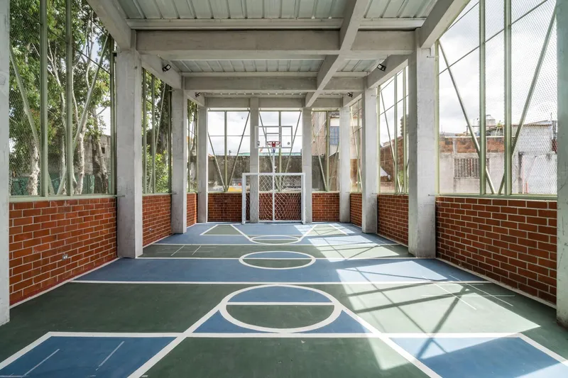 Indoor basketball court with red brick half-walls, concrete columns, exposed beams, and mesh-screened diagonal-braced windows casting angular shadows