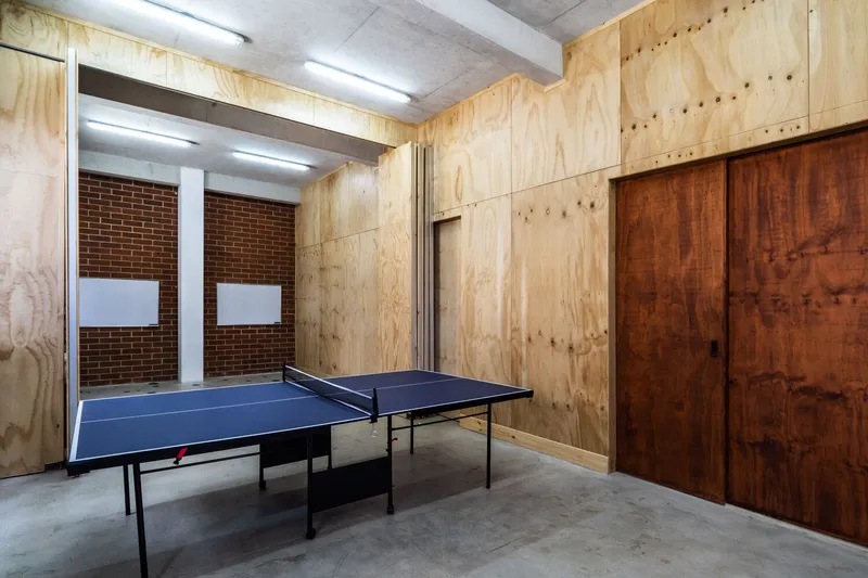 Recreation room with blue ping-pong table, plywood wall panels, exposed concrete ceiling beams, and red brick window wall