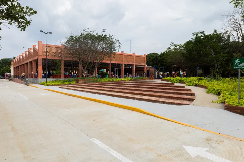 Paved plaza with curved timber steps and planted beds leading to perforated brick pavilion under cloudy skies