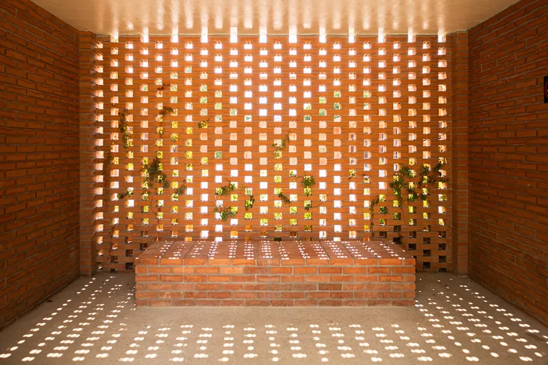 Perforated brick wall with grid openings filtering daylight onto brick seat and greenery beyond
