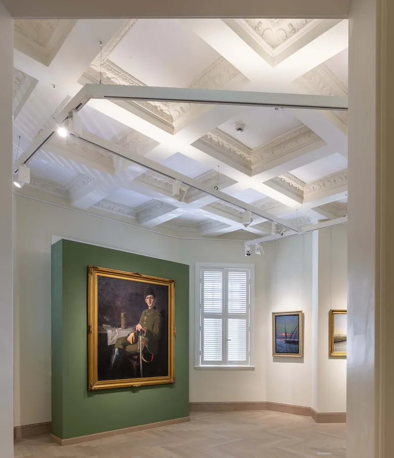 Gallery room with coffered ceiling, green partition wall and framed artworks under track lighting