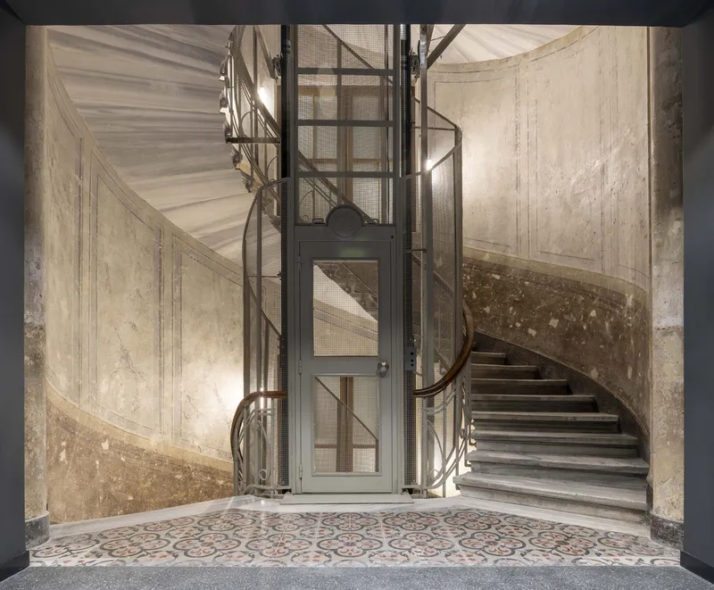 Curved stone staircase with steel and glass elevator shaft and patterned tile floor