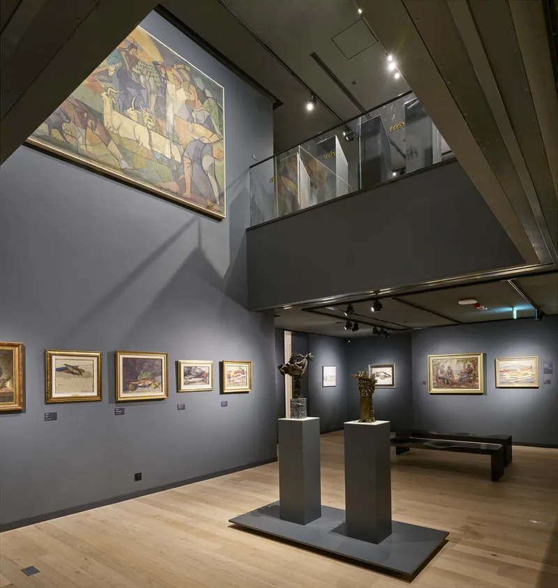 Double-height gallery space with grey walls, glass balustrade mezzanine, and timber floor with sculptures
