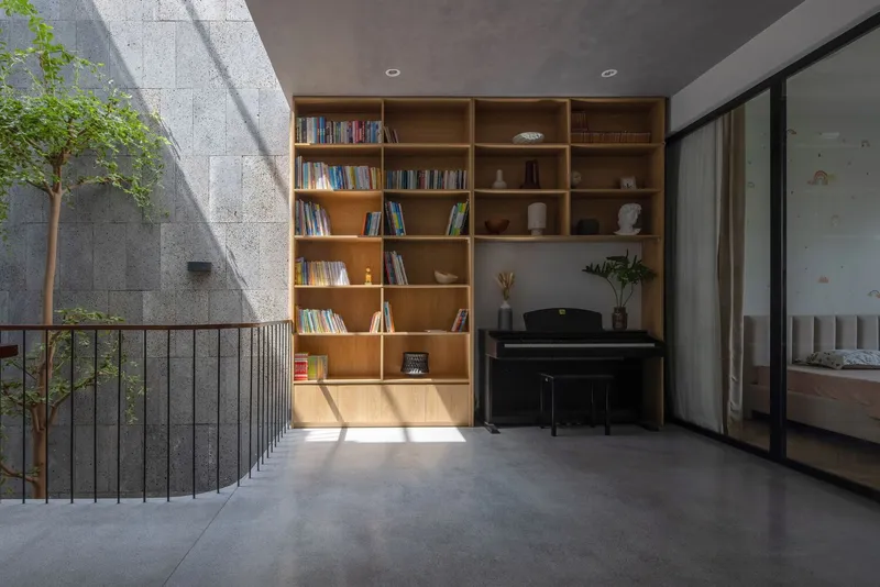Mezzanine alcove with timber bookshelf and piano receiving sharp afternoon light from the courtyard void