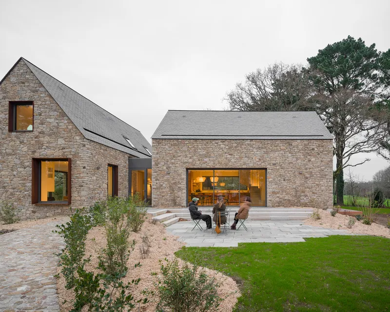 Courtyard terrace with stone paving and chairs beneath two gabled stone volumes at dusk