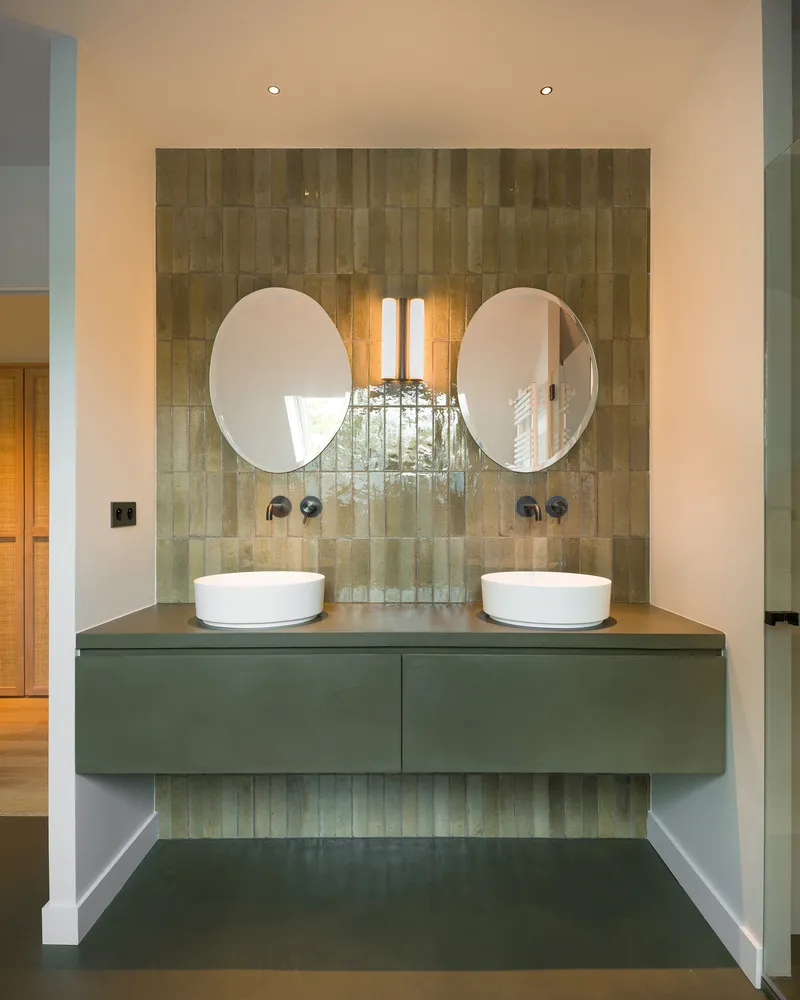 Bathroom vanity with twin vessel sinks, circular mirrors and vertical glazed tile wall under recessed lighting